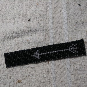 May include: Black beaded bracelet with a white arrow design. The arrow is centered on the bracelet and is made of small white beads. The bracelet is rectangular and appears to be handmade.