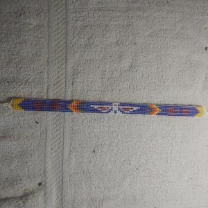 May include: A beaded bracelet with a blue background and yellow, red, and white accents. The bracelet has a geometric pattern.