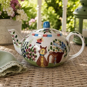 May include: A ceramic teapot with a crackle glaze finish featuring hand-painted garden motifs including a brown cat, a birdhouse, a welcome sign, and floral patterns. The teapot has a blue lid knob, a curved handle, and a spout with painted flowers.