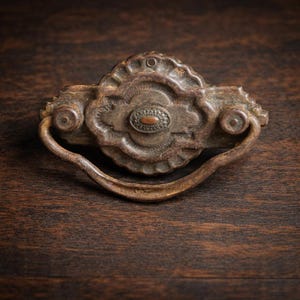 May include: Antique bronze drawer pull with a decorative oval center and curved handle. The hardware has a distressed finish, showing signs of age and wear. The pull is mounted on a dark wood surface.