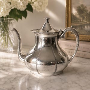 May include: A polished silver teapot featuring a rounded body with vertical panels decorated with a Greek key pattern. The teapot has a curved spout, a matching lid with a small finial knob, and a curved handle with ribbed detailing.