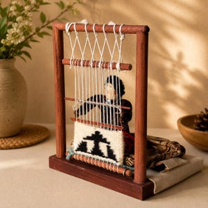 May include: A wooden loom frame with a small doll positioned behind the warp threads. The doll is weaving a white textile featuring a black geometric pattern. The loom is constructed from dark brown wood and includes white string warp threads.
