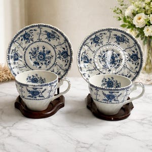 May include: Two matching teacups and saucers with a blue floral design on a white background. The teacups and saucers are displayed on dark wooden stands. The teacups have a curved handle and a detailed blue floral pattern. The saucers have a scalloped edge.