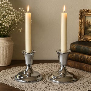 May include: Two polished silver-toned metal candle holders with flared bases and rims, each supporting a single lit cream-colored taper candle. The holders feature a smooth, reflective surface and a classic pedestal design, standing upright.