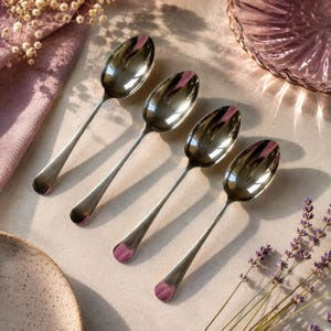 May include: Four silver spoons with a pink accent on the handle tips are arranged diagonally on a light-colored surface. The spoons are reflective, and the scene includes a pink napkin, a decorative plate, and sprigs of lavender.