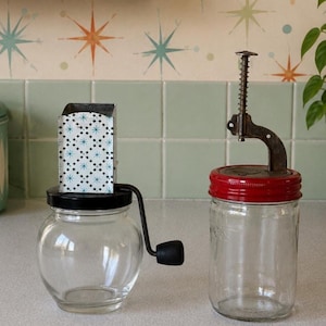 May include: A clear glass jar with a black lid, a hand crank, and a white metal housing featuring a blue star pattern, alongside a clear glass jar with a red lid and a vertical metal spring-loaded mechanism attached to the top.