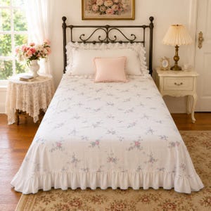 May include: A white bedspread featuring a delicate blue and pink floral pattern with a grid-like texture and a ruffled bottom edge. The bed is styled with two white ruffled pillows and a single solid light pink square accent pillow.