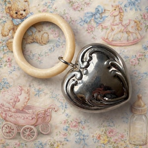May include: A baby rattle featuring a light-colored wooden ring and a silver heart-shaped charm. The heart has an ornate, embossed design. The background includes illustrations of a teddy bear, a rocking horse, a baby carriage, and a baby bottle.