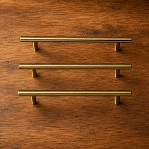 May include: Three cylindrical cabinet pulls with a brushed brass finish. Each handle features a long, straight bar supported by two perpendicular posts at either end, creating a clean, linear design for furniture or cabinetry hardware.