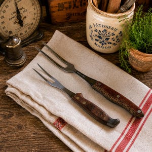 May include: Two vintage meat forks with wooden handles rest on a linen towel with red stripes. The forks have a stainless steel appearance and are positioned diagonally. The background includes a scale, a ceramic jar, and a small wooden box.