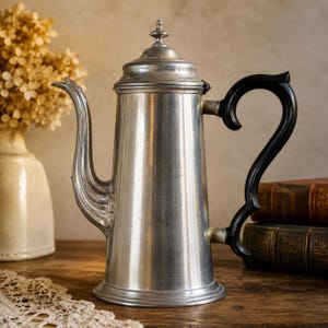 May include: A tall, cylindrical metal coffee pot with a polished silver-toned finish, featuring a curved spout, a matching lid with a small finial top, and a dark, curved handle. The vessel has a wide base and a smooth, vertical body design.