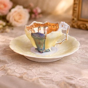 May include: A vintage teacup and saucer set. The teacup has a yellow exterior with gold trim and a decorative handle. The interior of the cup is gold. The saucer is a matching pale yellow. The set is placed on a lace tablecloth.