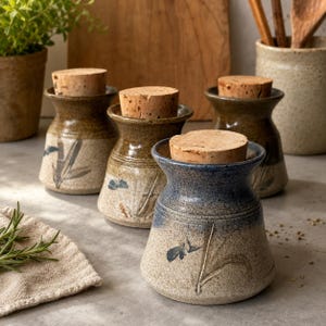 May include: Three ceramic jars with cork stoppers, each featuring a flared rim and a speckled beige base with hand-painted botanical motifs. The jars have a gradient glaze transitioning from blue or brown at the top to a speckled cream at the bottom.