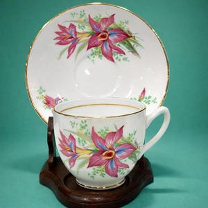 May include: A white teacup and saucer set with a floral design. The cup and saucer have a gold trim and a pattern of pink and blue flowers with green leaves. The set is displayed on a dark wooden stand.