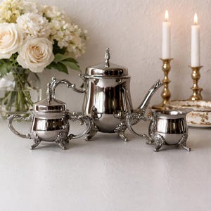 May include: A polished silver tea set with ornate handles and feet. The set includes a teapot, sugar bowl, and creamer. The teapot has a lid with a decorative finial. The set is displayed with white roses in a glass vase and lit candles.