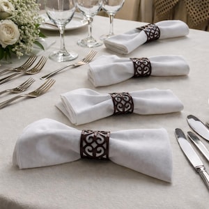 May include: White cloth napkins are held by decorative brown metal napkin rings. The rings have an openwork design. Silver forks, knives, and wine glasses are also on the table, along with a small bouquet of white flowers.