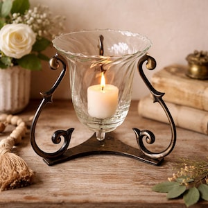 May include: A decorative candle holder with a lit white candle inside a clear glass hurricane. The glass is supported by a bronze-colored metal base with ornate, curved arms. The candle holder sits on a wooden surface.