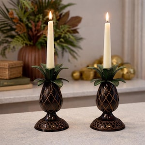 May include: Two decorative pineapple-shaped candle holders with lit ivory candles. The candle holders are dark brown with green leaf accents and a textured design. The candles are approximately 10 inches tall.