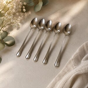 May include: A set of five silver-toned metal spoons arranged in a row. Each spoon features a smooth, polished oval bowl and a slender handle with a subtle, raised decorative border detail along the edges, terminating in a rounded end.