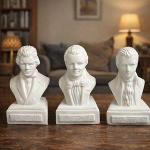 May include: Three white ceramic busts of famous composers: Beethoven, Schubert, and Mozart. Each bust is mounted on a rectangular base with the composer's name and birth/death years. The busts are displayed on a wooden table.
