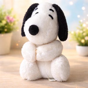 May include: A plush Snoopy toy, featuring a white body, black ears, and a black nose. The toy is in a seated position with its paws clasped together. The background is softly blurred with hints of greenery.