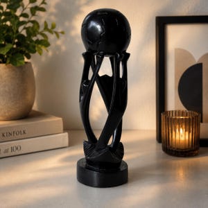 May include: A black sculpture featuring three stylized figures standing on a circular base, their arms reaching upward to support a smooth, spherical globe. The piece has a polished, dark finish and a vertical, elongated design.
