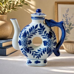 May include: A white and blue ceramic teapot with a unique circular design. The teapot features a blue floral pattern and a curved handle. The spout and lid are white, and the handle is a deep blue. A decorative piece for the kitchen or dining room.