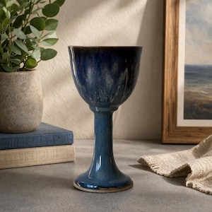 May include: A tall, blue ceramic goblet with a flared rim and a slender stem. The goblet has a glossy finish with darker blue streaks and a brown rim. It is displayed on a gray surface, next to a stack of books and a framed picture.