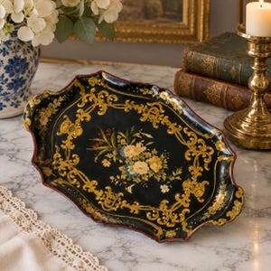 May include: An oval black metal tray featuring a central floral bouquet of yellow and blue flowers, surrounded by intricate gold scrollwork and filigree patterns along the scalloped edges, with a thin red painted border trim.