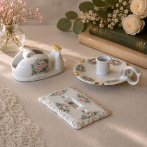 May include: A white porcelain tape dispenser, a candle holder with a handle, and a rectangular light switch cover plate. Each item features a hand-painted pink rose floral pattern with green leaves and gold-painted scalloped edges.