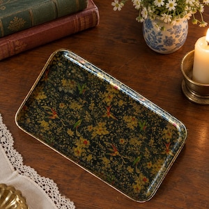May include: A rectangular serving tray with a black base featuring a dense, intricate pattern of gold-toned flowers, foliage, and small birds. The tray has a metallic gold-colored rim and a smooth, reflective surface finish.
