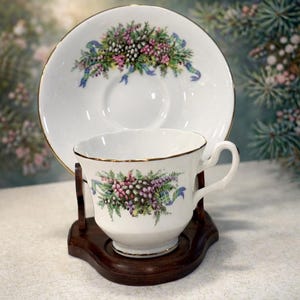 May include: White teacup and saucer set with a floral design in pink, purple, and green, accented with blue ribbons. The cup and saucer have gold trim. The set is displayed on a dark wooden stand, showcasing a classic tea set.