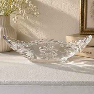 May include: Clear glass serving dish with a starburst design in the center. The dish has a scalloped edge and is shaped like a boat. A vase with white flowers and a framed picture are in the background.