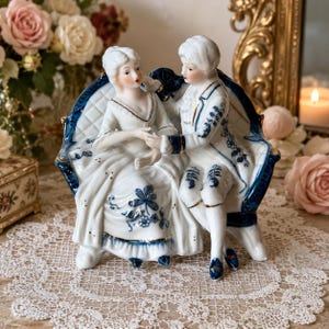 May include: A porcelain figurine of two figures sitting on a curved bench, decorated with white and blue floral patterns and gold trim. The figures are dressed in period clothing, with the woman in a gown and the man in a coat and breeches.