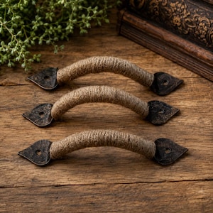 May include: Three drawer pulls featuring a curved metal handle wrapped in natural tan jute rope. Each end of the handle is secured by a dark, textured metal mounting plate shaped like a pointed spade with a single pre-drilled screw hole.
