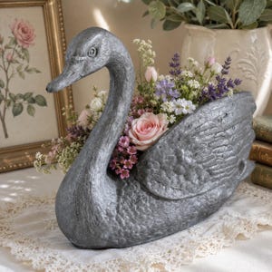 May include: A silver-colored swan planter filled with a variety of flowers, including pink roses, lavender, and white blossoms. The swan has a textured surface and is sitting on a lace doily. A framed picture is in the background.