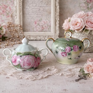 May include: Two antique porcelain sugar bowls with lids. One is pale blue with pink roses, the other is green with pink flowers and gold trim. Both have handles and sit on a lace doily, with a bouquet of pink roses in the background.