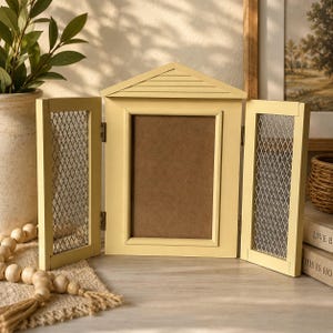 May include: A pale yellow wooden picture frame shaped like a house with a peaked roof. The frame features two hinged side doors with silver-toned metal mesh panels and a central rectangular opening for a photograph, all finished in a matte paint.