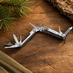 May include: A silver-toned Winchester multi-tool featuring several extended implements including a nail clipper, a small blade, and a file. The metal tool is hinged and includes a small circular ring at one end for attachment, with the brand name clearly engraved on the side of the main body.