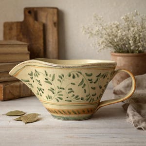 May include: Cream-colored ceramic gravy boat with a handle and spout. The boat is decorated with green leaf patterns and horizontal bands of orange and green. The design has a rustic, vintage aesthetic.