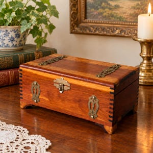 May include: A rectangular wooden box with a warm brown finish, featuring decorative brass-toned metal plates on the lid and front. The box has a central metal latch, dark-toned finger joint corners, and four small feet at the base.