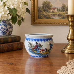 May include: A white ceramic planter with a rounded body, featuring a blue heart-patterned border at the rim and a colorful illustration of figures in traditional attire on a boat, with a gold-toned trim along the top edge.