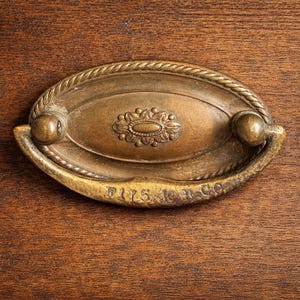 May include: Antique brass drawer pull with an oval design and rope-like edging. The center features an ornate floral pattern. The text "F175 L & Co" is visible at the bottom. The pull is mounted on a wooden surface.