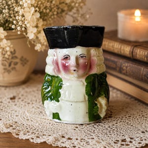 May include: A ceramic character jug shaped like a person wearing a black tricorn hat, a white powdered wig, and a green coat over a white shirt. The figure has rosy cheeks, a small mouth, and a glossy finish, depicting a historical style figure.