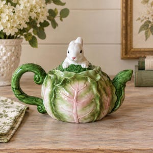 May include: A ceramic teapot shaped like a head of cabbage with green and pink leaves. The lid features a sculpted white rabbit sitting atop the cabbage. The teapot has a curved green handle and a short spout, with a glossy finish throughout.