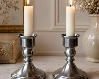 May include: Two matching silver-toned metal candle holders with a smooth, polished finish. Each holder features a wide, circular base, a slender stem, and a cylindrical top cup. A single, tall, off-white pillar candle is lit in each holder.