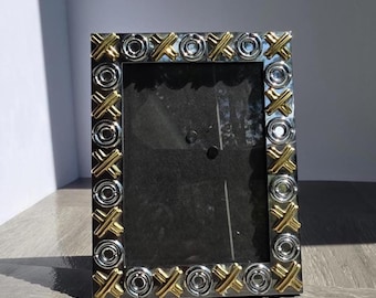 May include: A rectangular photo frame with a silver and gold trim. The frame has a repeating pattern of gold X's and O's around the border. The frame is empty, with a dark background visible through the glass.