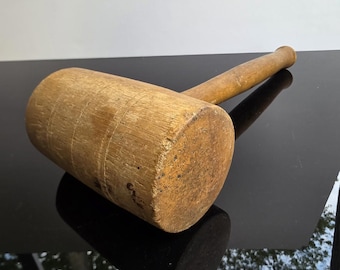 May include: A wooden mallet with a cylindrical head and a long, rounded handle. The mallet is made of light-colored wood, showing visible grain and wear. The handle is attached to the head, creating a tool for striking or shaping materials.