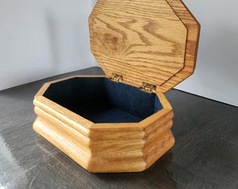 May include: An open, wooden jewelry box with an octagonal shape. The box is made of light-colored wood with a visible grain. The interior is lined with dark blue fabric. The lid is partially open, revealing the interior.