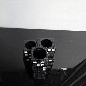 May include: A black ceramic vase with a cityscape design. The vase has three cylindrical openings and is decorated with white rectangular accents to represent windows. The vase is on a reflective black surface.